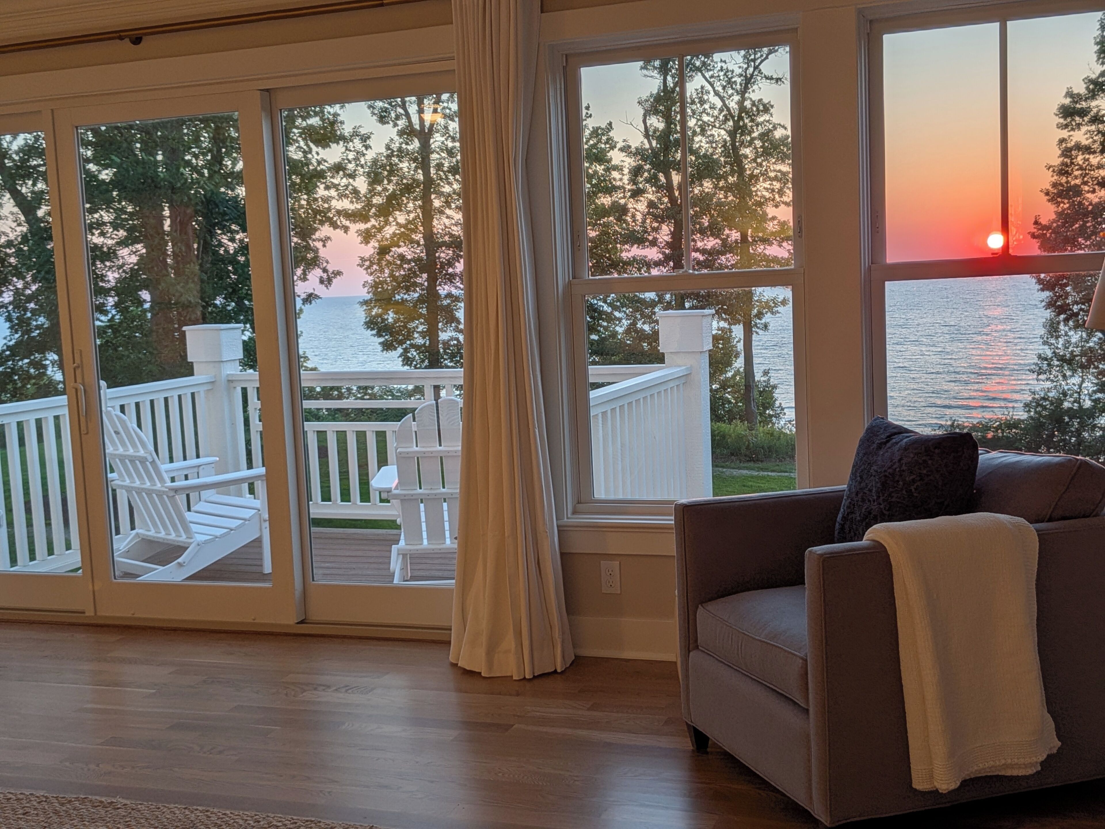 Une pièce dotée de grandes fenêtres donnant sur une terrasse et l’eau, avec un canapé et une vue sur le coucher de soleil.