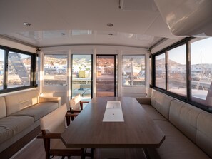 A modern interior with a long wooden table, cushioned seating, and large windows offering views of the outside. - Sun-Kissed Sailing Adventure in the Stunning Islas Cícladas (Paros)