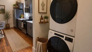 A compact kitchen with modern appliances, a dining area with a chair, and a laundry area with stacked washers and dryers. - Short Walk to Track & Broadway, SPAC Close Too! (Saratoga Springs)