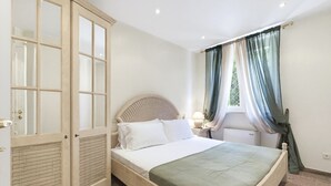Un dormitorio con cama, armario, ventana con cortinas y mesita de noche con lámpara.