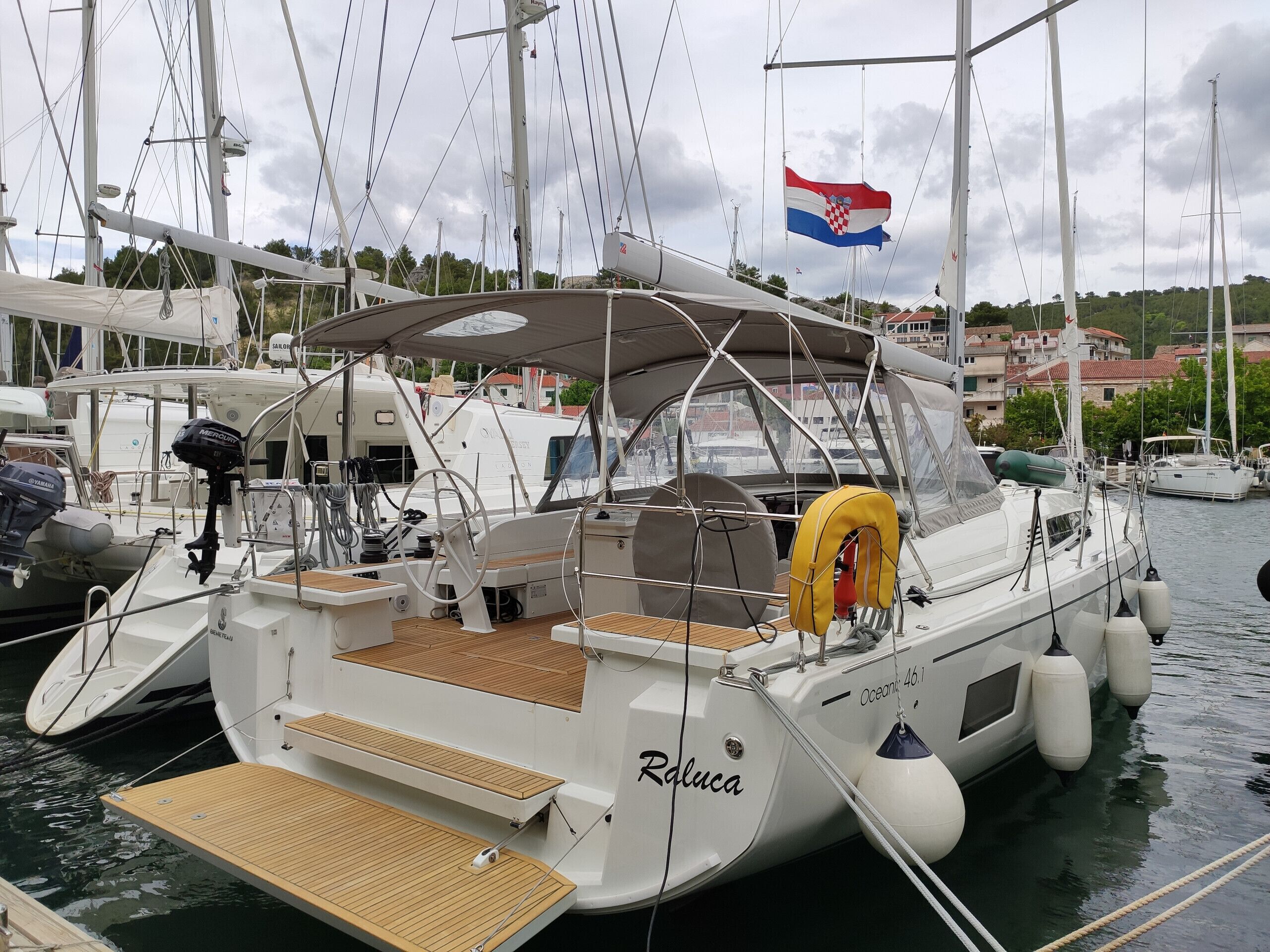 A sailboat docked at a marina with a flag, a steering wheel, and a name 'Raluco' on the side.