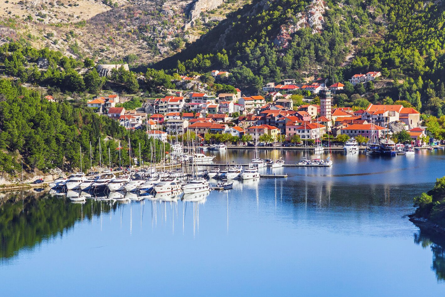 A coastal town with a marina, houses with red roofs, and a mountainous backdrop.