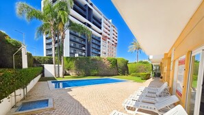 Uma área de piscina com espreguiçadeiras, uma piscina pequena e um edifício com varandas.