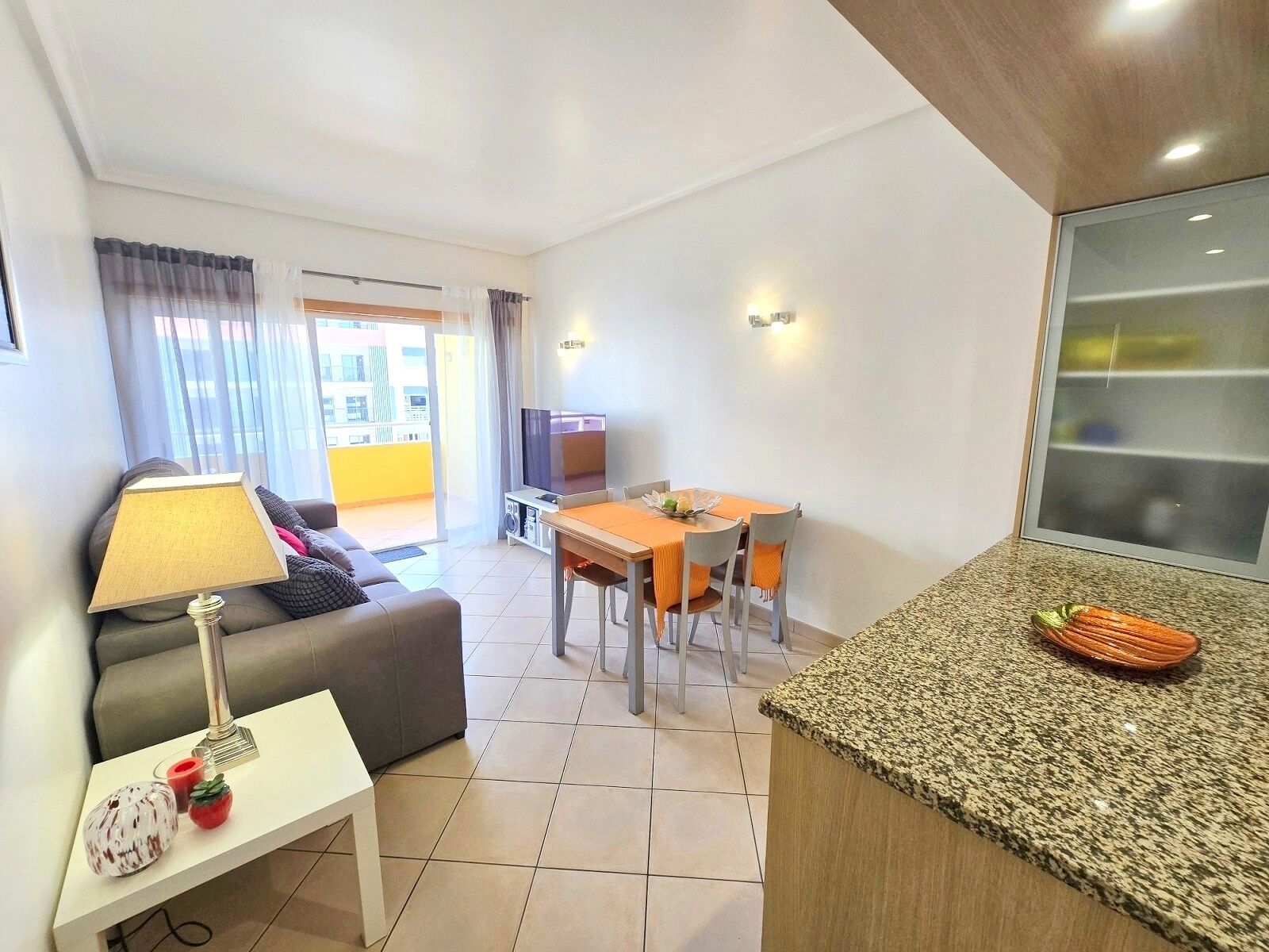 Una sala de estar moderna con sofá, mesa de comedor, televisor y zona de cocina con encimera de granito.
