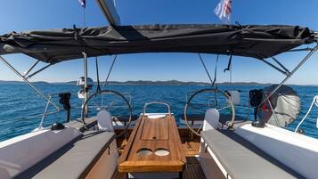 A boat deck with a wooden table, two white cushioned seats, a steering wheel, and a flag.