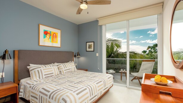 A bedroom with a bed, a ceiling fan, a window with a view, a nightstand, and a bedside table. - Rooftop Penthouse in Akumal jungle — steps to the beach (AKUMAL)