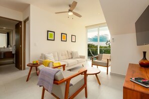 Een moderne woonkamer met een bank, stoelen, een salontafel en een televisie. Er is een balkon met uitzicht op de lucht en groen.