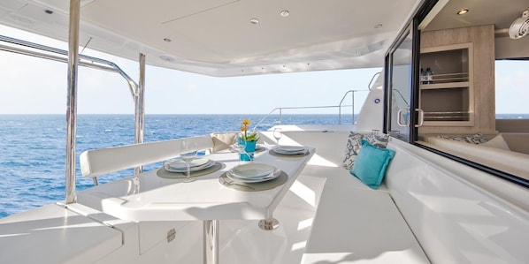 A dining area on a boat with a table set for two, a view of the sea, and a cabin with a window.