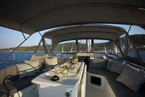 A boat interior with a dining area, cushioned seating, and a view of the water and hills. - Sun-Kissed Sailing Adventure in Islas Sarónicas (Alimos)