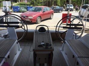 A boat's deck with two steering wheels, a small table with a bowl, and a view of parked cars and people in the background. - Sun-Kissed Sailing Adventure in Islas Esporadas (Skiathos)