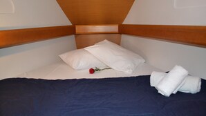 Un lit simple avec une tête de lit en bois, des oreillers blancs, une couvre-lit bleu foncé, une rose rouge et des serviettes blanches pliées.
