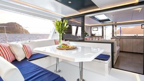 Ein modernes Bootsinnere mit Essbereich, Sitzgelegenheiten mit Kissen, einem Tisch mit Obstplatte und Blick aufs Wasser.