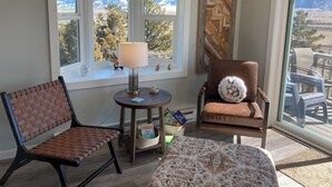 A corner room with a view of mountains, a lamp, a chair with a cushion, and a small table with books.