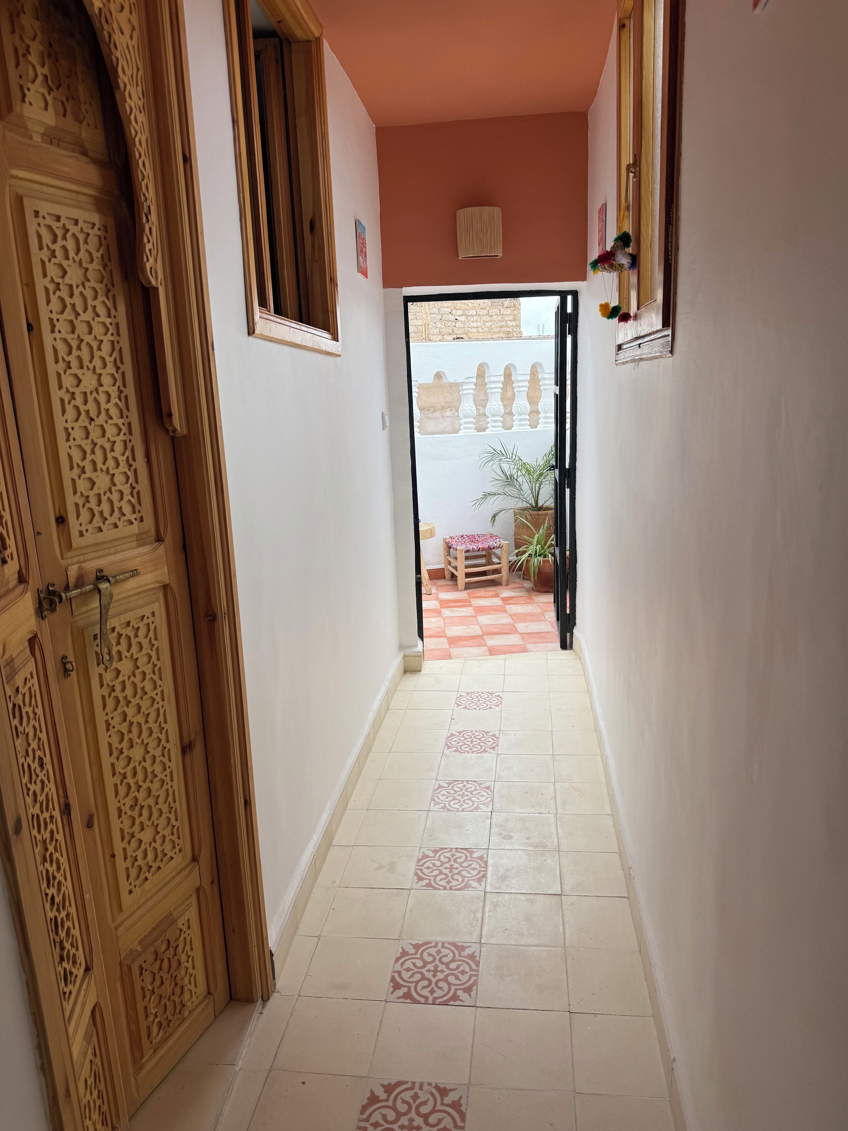 A hallway with patterned tiles, a wooden door with intricate carvings, and a small table with a potted plant.