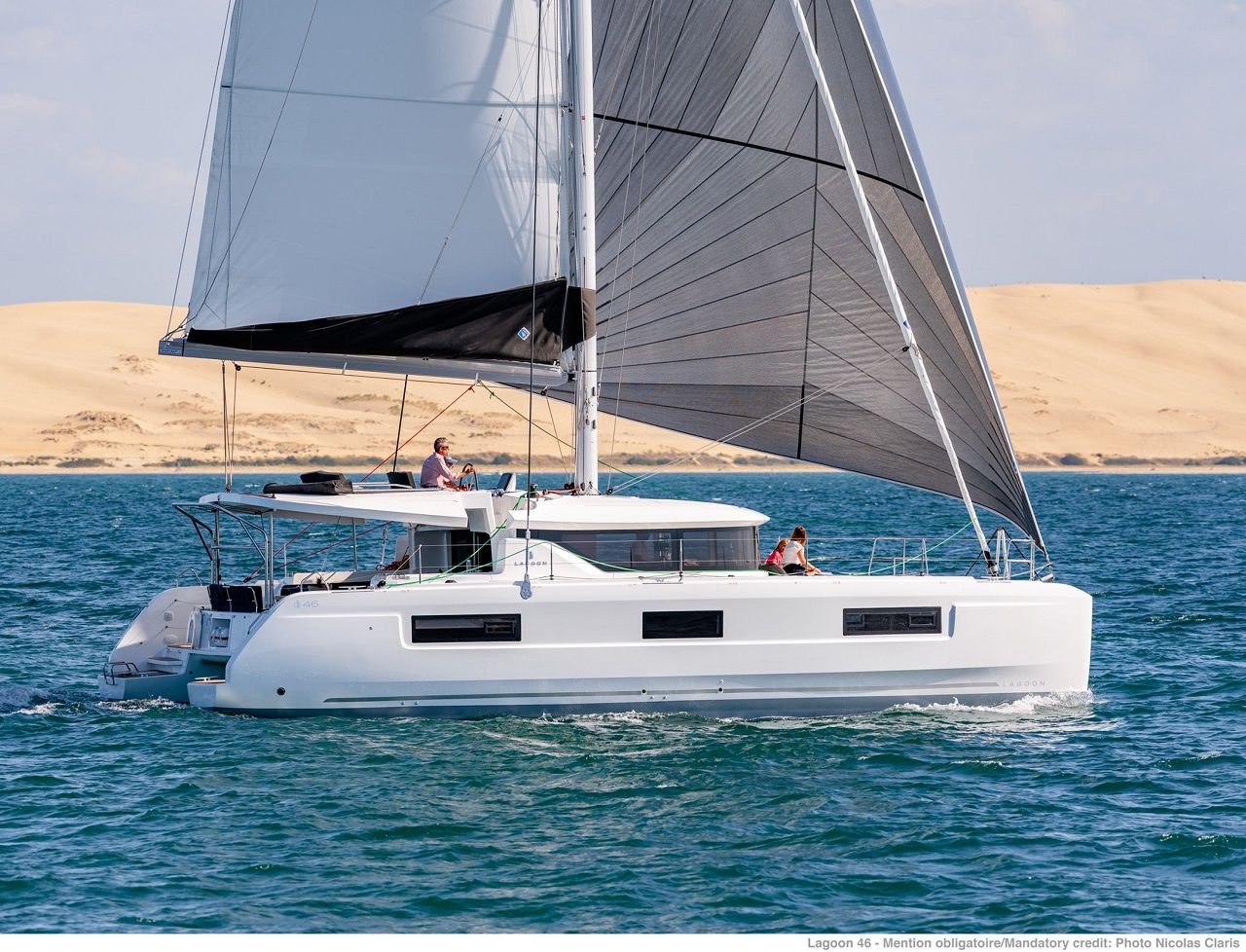 A white catamaran with a cabin and a sail, sailing on calm waters with a desert landscape in the background.