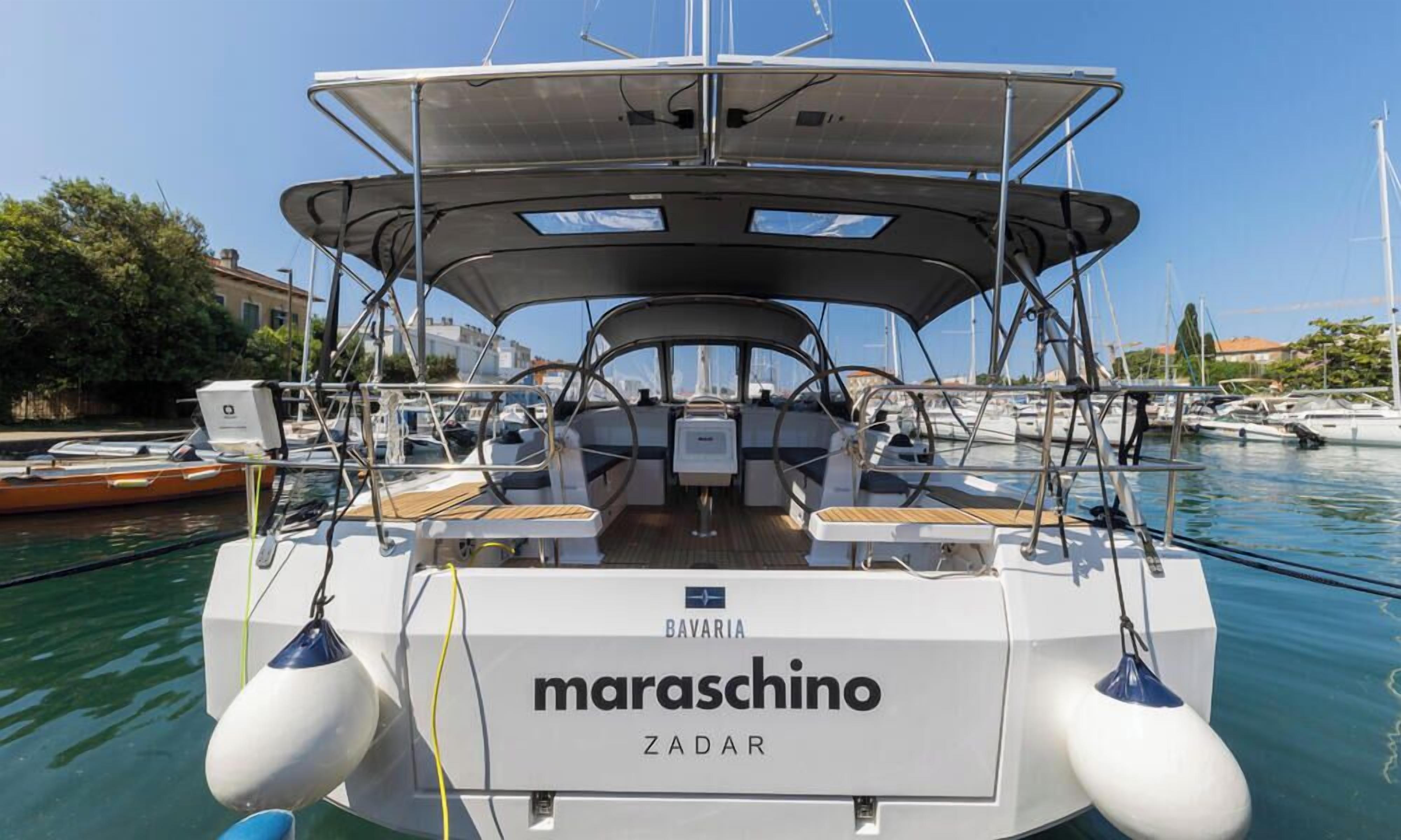 A boat named 'BAVARIA' with a 'maraschino' and 'ZADAR' branding, docked at a marina with other boats and buildings in the background.