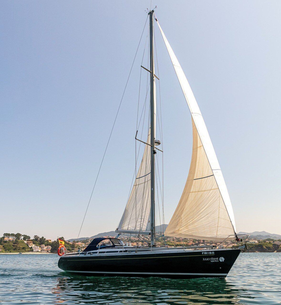 A sailboat with a tall mast and a single sail, anchored in calm waters with a coastal town in the background.