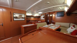 A dining area with a table set for two, a television mounted on the wall, and a kitchen area in the background. - Sun-Kissed Sailing Adventure in Islas Sarónicas (Alimos)