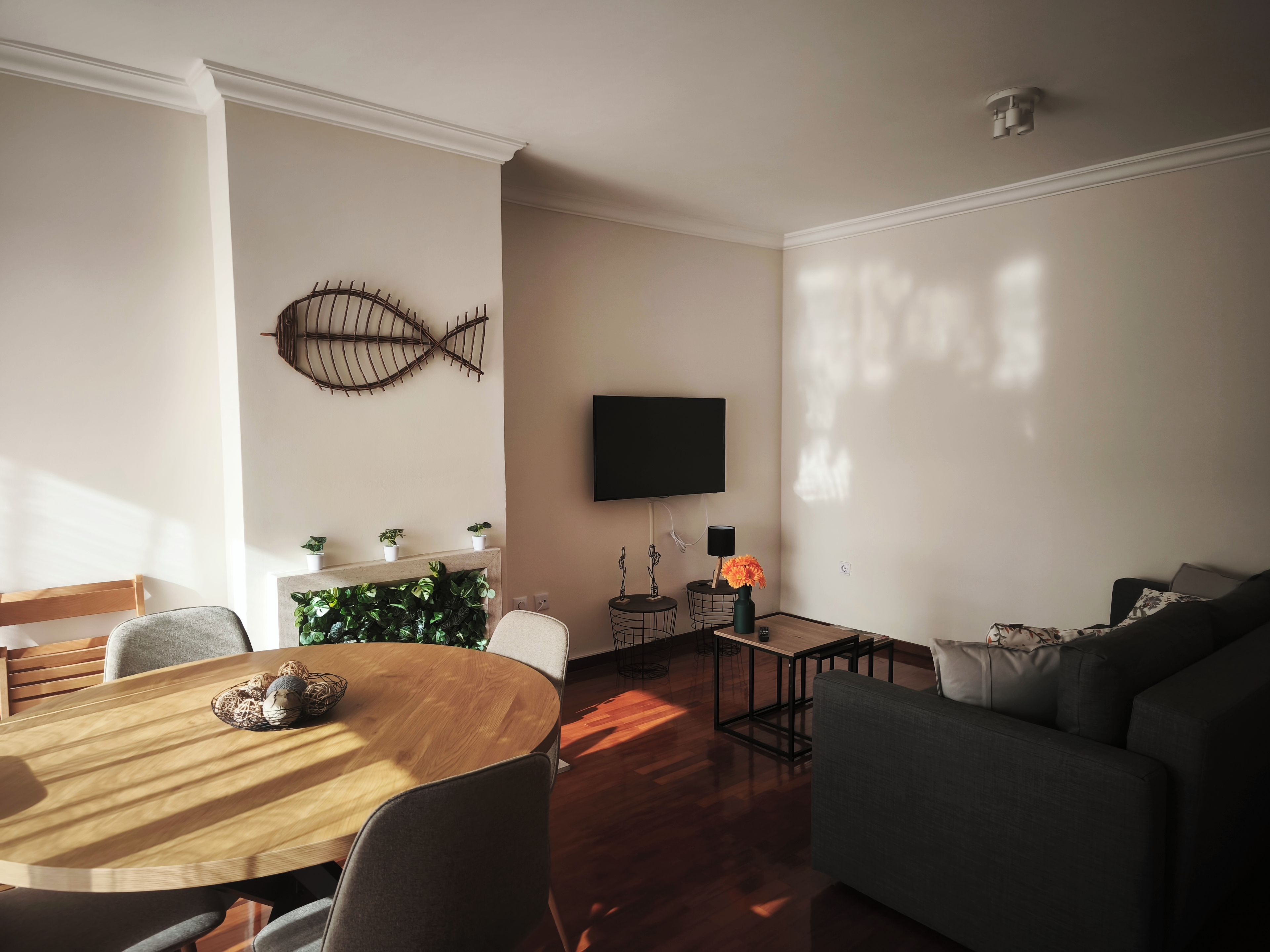 A living room with a round wooden table, a sofa, a TV mounted on the wall, and a fish-shaped wall decoration.