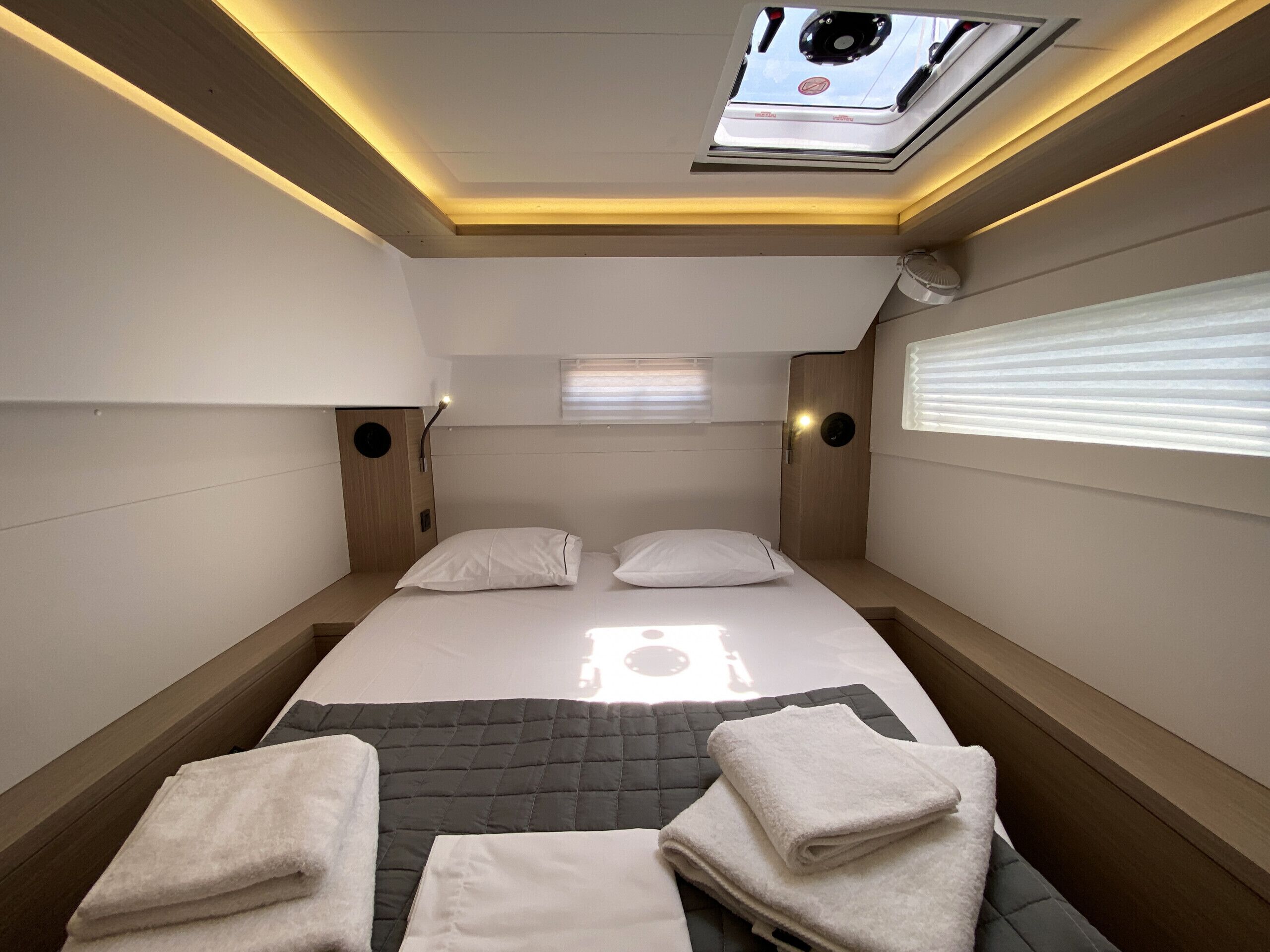A compact hotel room with a bed, two pillows, a bedside table, a window with blinds, and a skylight.