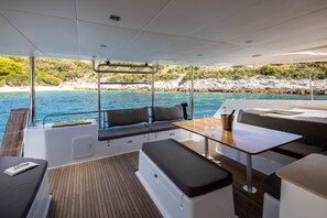 Interior de un barco con asientos, una mesa y vistas al mar y a la costa rocosa.