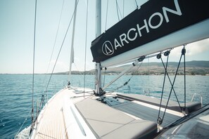 Eine Segelyacht mit dem Schriftzug 'ARCHON' auf dem Segel, einem Deck mit Geländer und Blick auf das Meer und das entfernte Land.