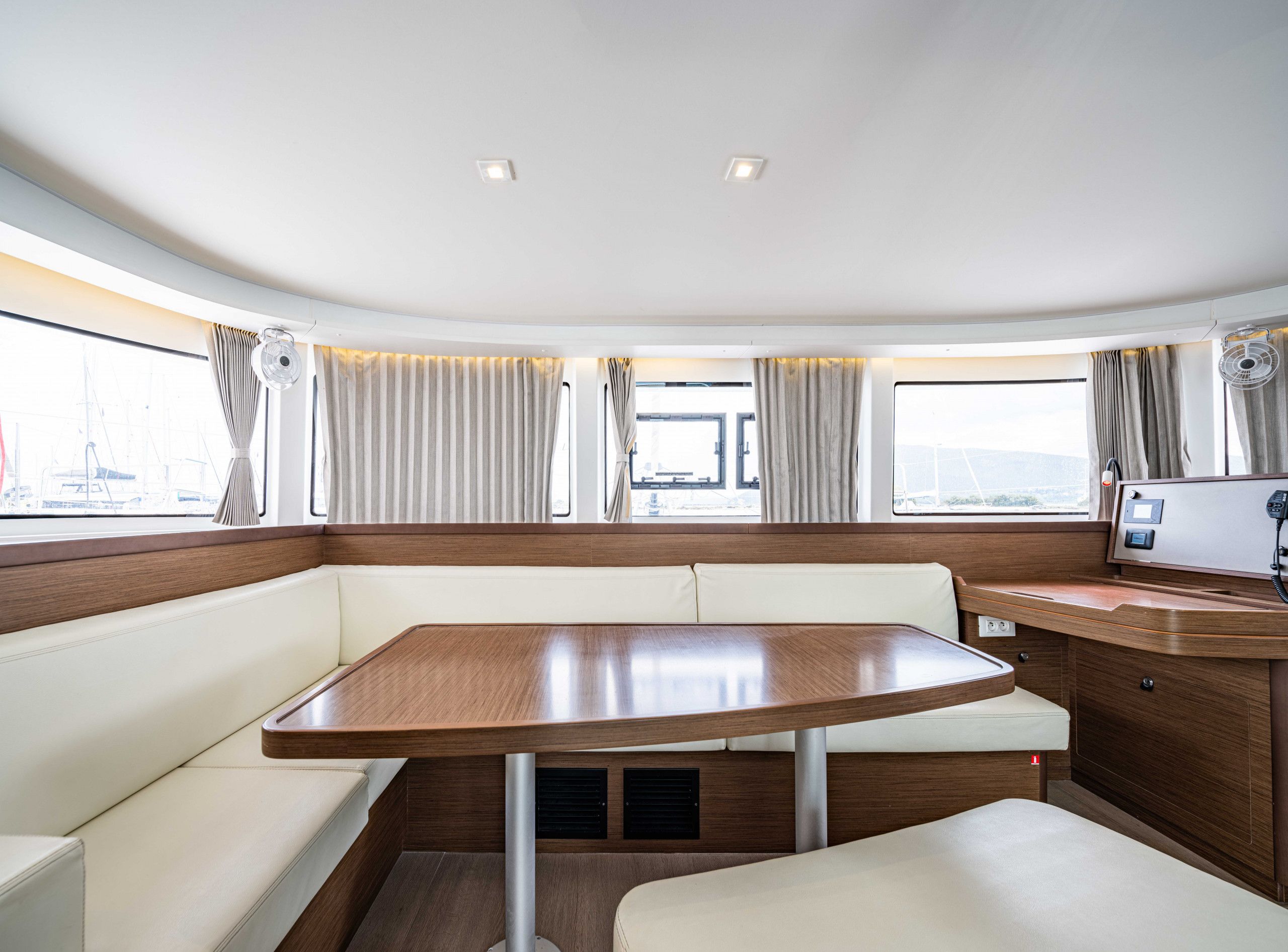 A cabin with a wooden table, cushioned seating, a control panel, and large windows with curtains.