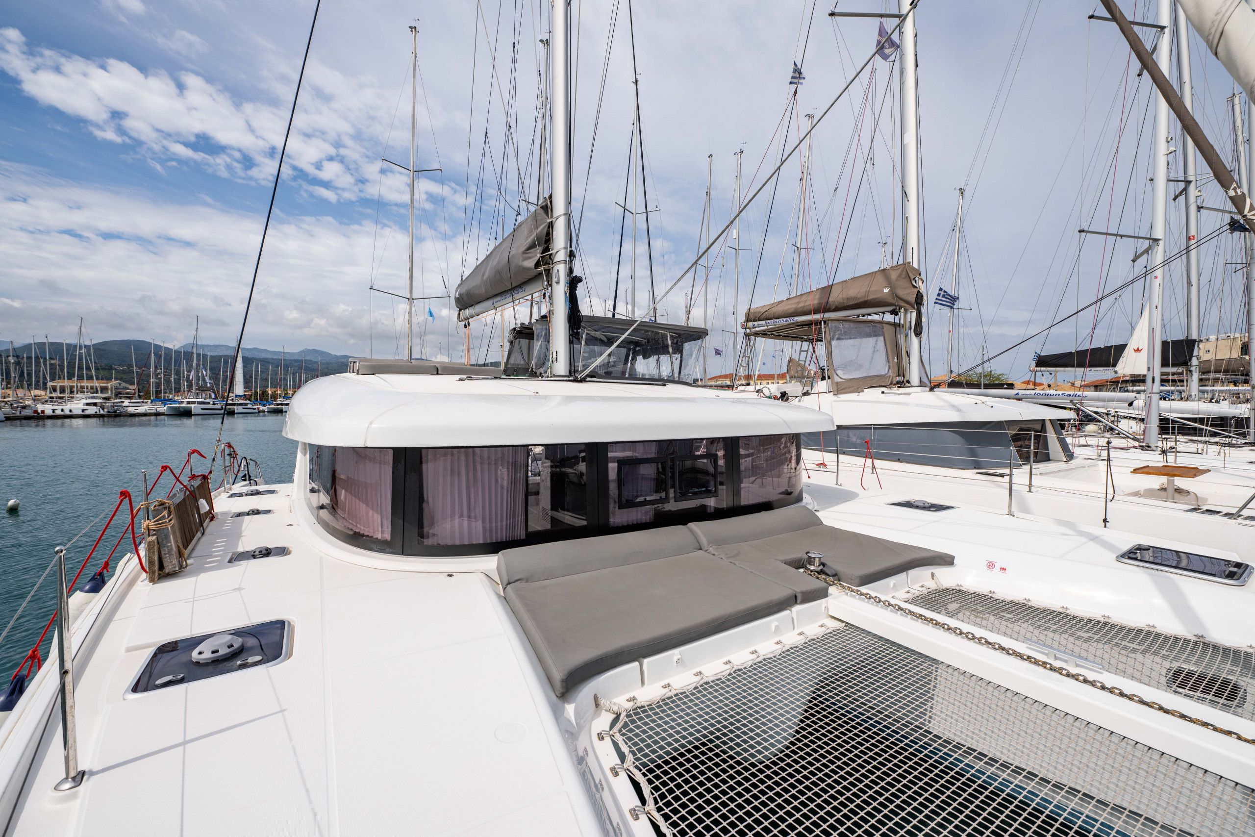 Un'imbarcazione a vela ormeggiata in una marina, con diversi alberi, una zona coperta per sedersi e vista su altre imbarcazioni e sulla costa in lontananza.