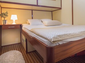 A neatly made bed with a patterned bedspread, a bedside lamp, a potted plant, and a small rug on a wooden floor. - Sun-Kissed Sailing Adventure in Split-Dalmacia, Croatia (Jesenice)