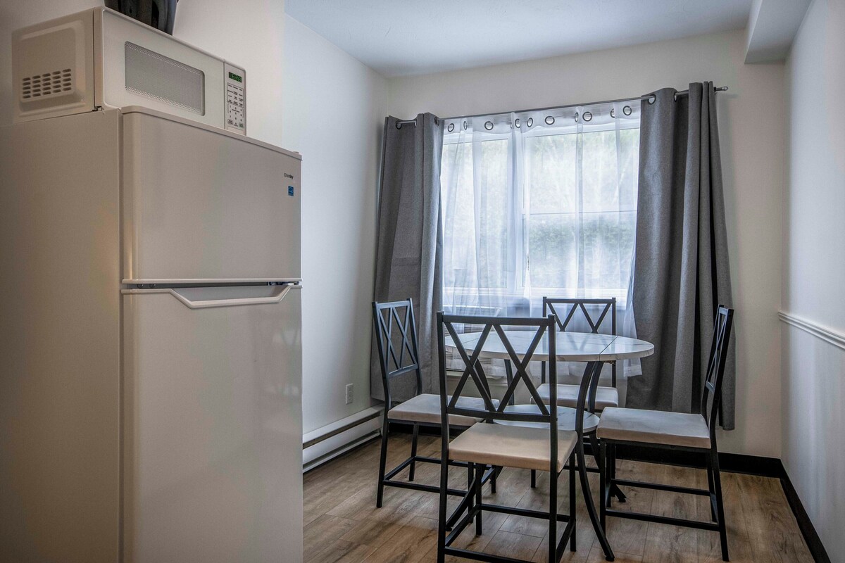 Una cocina compacta con un refrigerador blanco, una mesa de comedor con cuatro sillas y una ventana con cortinas.