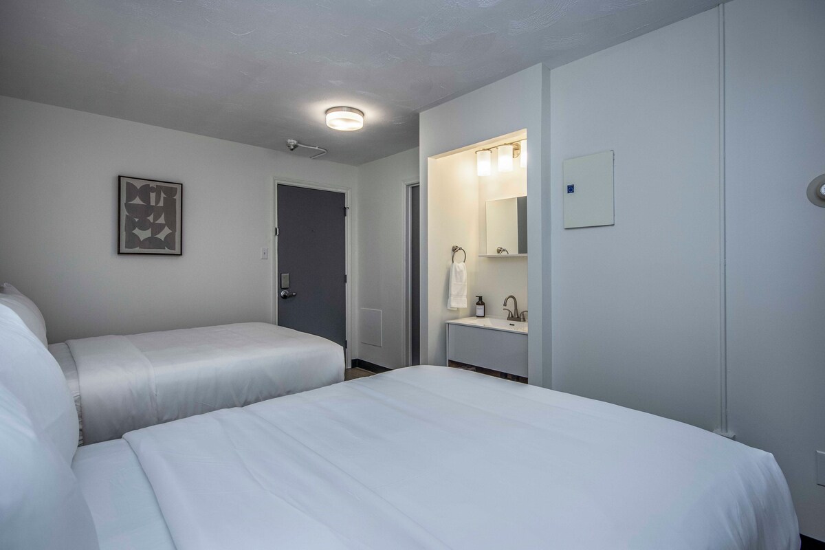 Habitación de hotel con dos camas, baño con espejo y lavabo, y puerta que da a otra habitación.