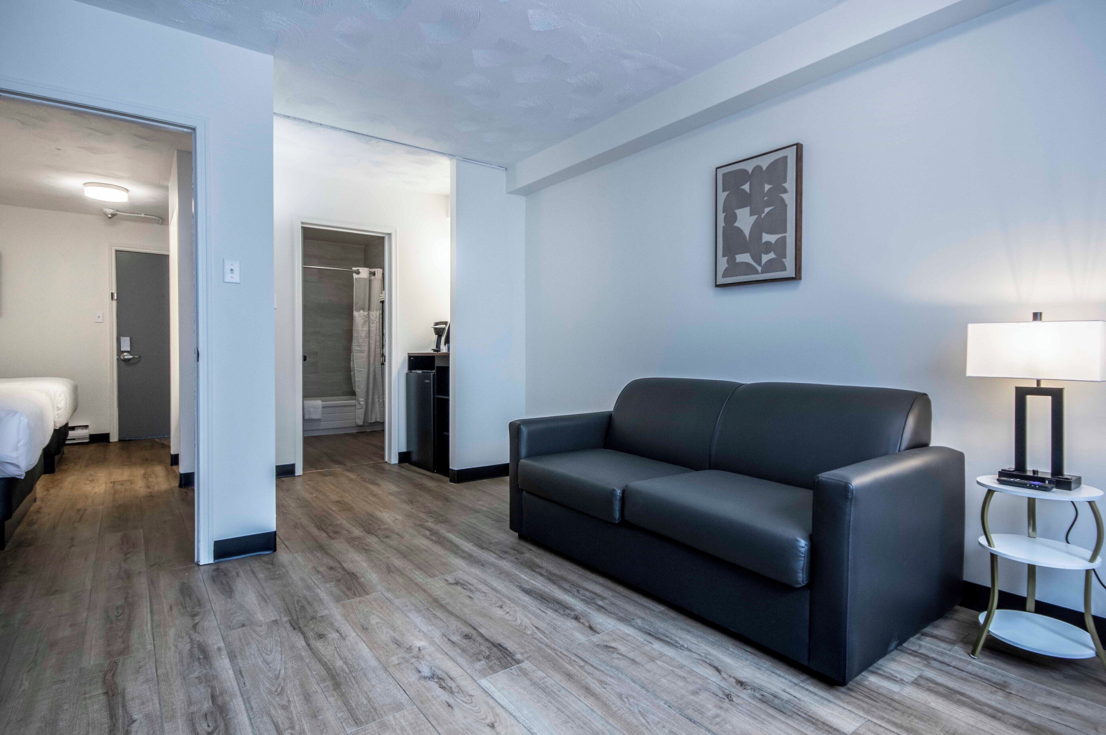 A modern hotel room with a grey leather sofa, a bedside table with a lamp, and a framed abstract artwork on the wall.