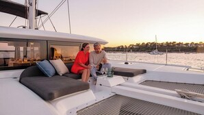 Et par sidder på dækket af en moderne yacht med udsigt over lystbådehavnen og solnedgangen.