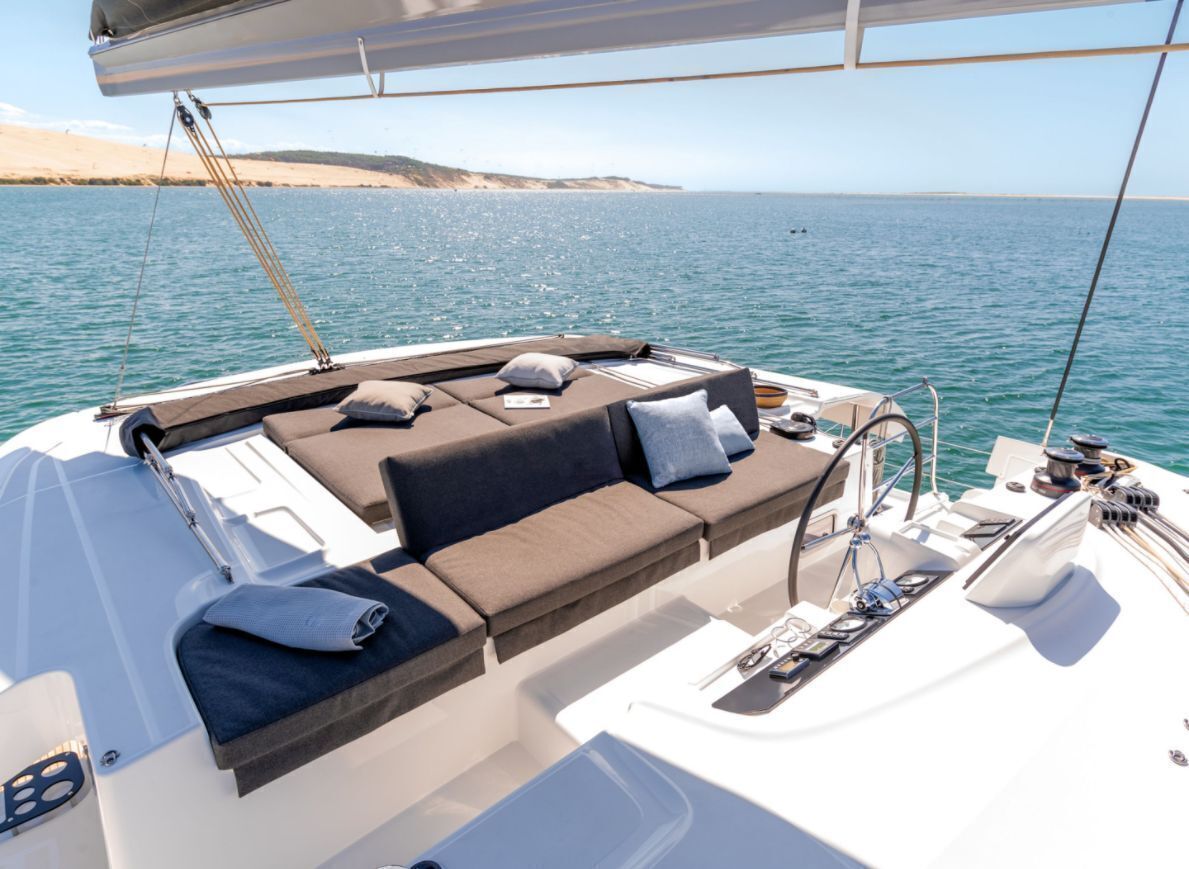 Cubierta de un velero con zona de asientos acolchada, una pequeña mesa y vistas al mar y la costa distante.