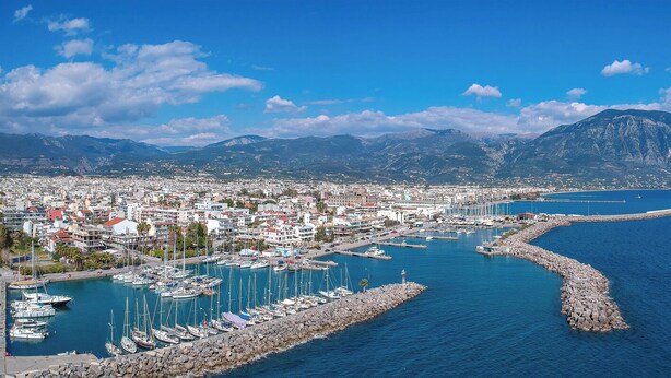 Eine Küstenstadt mit einem Yachthafen, einem steinernen Mole und Bergen im Hintergrund.