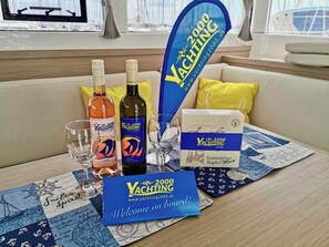A table with a welcome sign, two bottles of wine, glasses, and a box of crackers on a nautical-themed tablecloth.