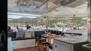 En yachts däck med sittplatser, ett bord och utsikt över hamnen och kullarna.