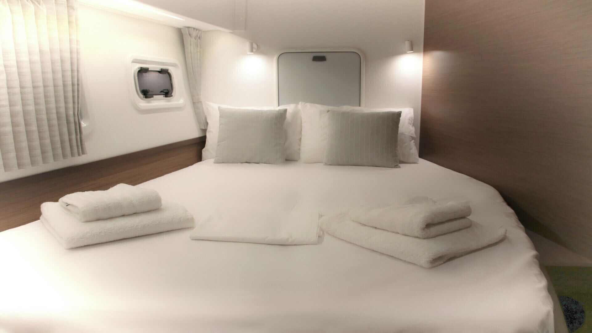 Ein ordentlich bezogenes Bett mit Kissen und gefalteten Handtüchern, ein kleines Fenster und eine Wandleuchte.