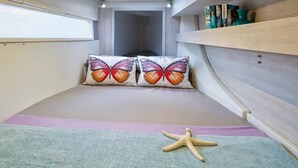 Une chambre à coucher de taille réduite, avec un lit superposé, deux coussins décorés de motifs de papillons, une étoile de mer en guise de décoration et une bibliothèque remplie de livres.