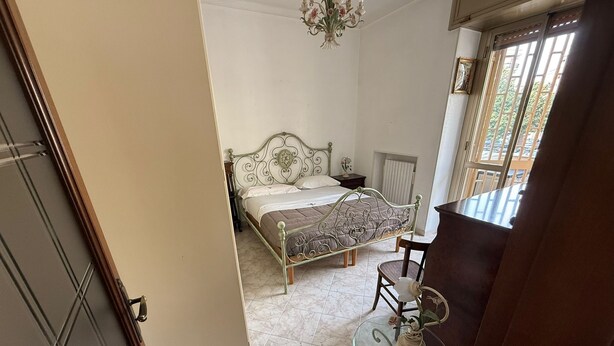 Une chambre à coucher avec un lit, une table en verre sur laquelle est posé un vase, et une fenêtre avec des stores.