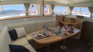 Interior de barco con sofá, mesa con comida y bebidas, y ventanas que dan a vistas del puerto deportivo.