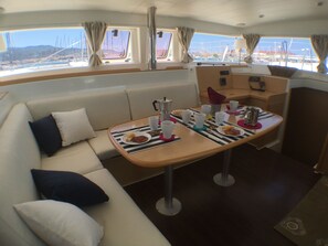 A boat interior with a sofa, table with food and drinks, and windows with views of the marina. - Sun-Kissed Sailing Adventure in the Ionian Islands, Lefkada (Lefkada)