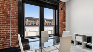 A modern room with a glass table, white chairs, a white shelving unit, and a view of buildings through large windows.