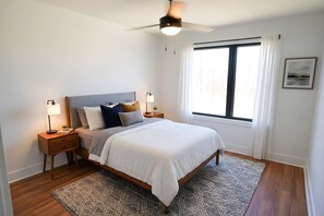 Ein Schlafzimmer mit einem Bett, Nachttischen, einem Deckenventilator, einem Fenster mit Vorhängen und einem gemusterten Teppich.