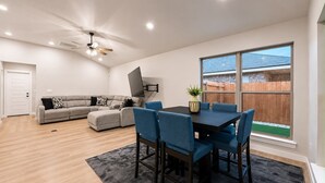 Ein modernes Wohnzimmer mit einer Eckcouch, einem Esstisch mit Stühlen, einem Deckenventilator und einem großen Fenster mit Blick auf einen Holzzaun.