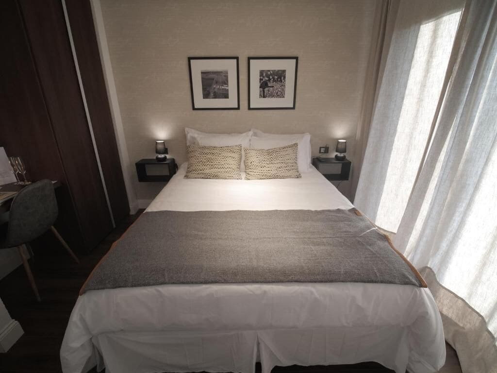 Uma cama bem arrumada com um cobertor cinzento, duas mesas de cabeceira com abajures e duas molduras com quadros na parede.