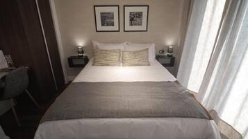 Uma cama bem arrumada com um cobertor cinzento, duas mesas de cabeceira com abajures e duas molduras com quadros na parede.