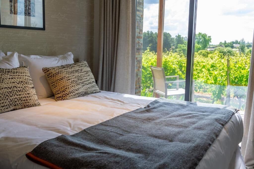 A bed with a grey blanket, white pillows, and a view of greenery through large windows.
