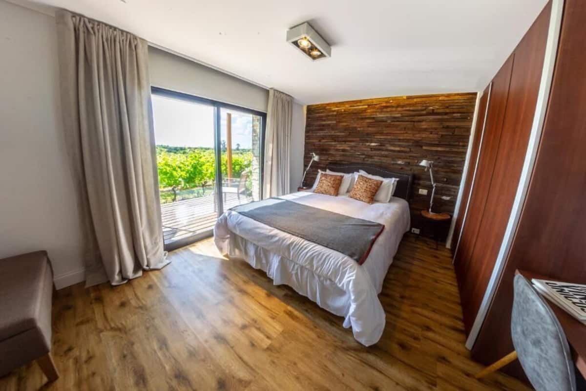 Un dormitorio con una cama grande, suelo de madera, vistas a través de una puerta corredera y una mesita de noche con una lámpara.