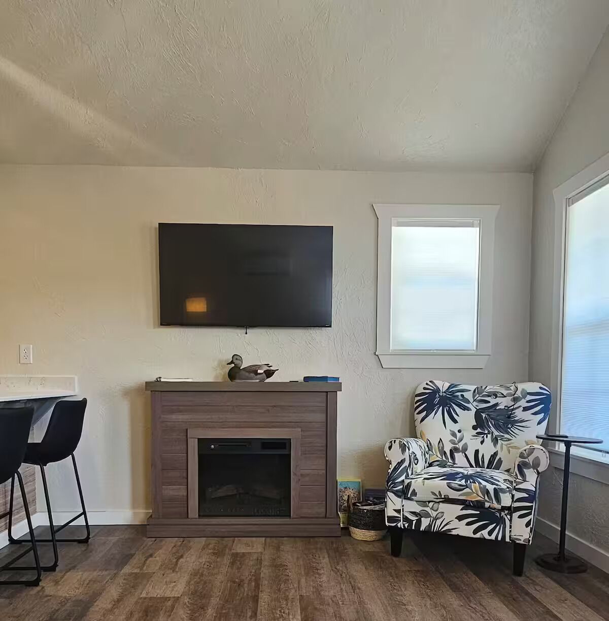 Una sala de estar con chimenea, un televisor montado arriba, un sillón estampado y una ventana con persianas.